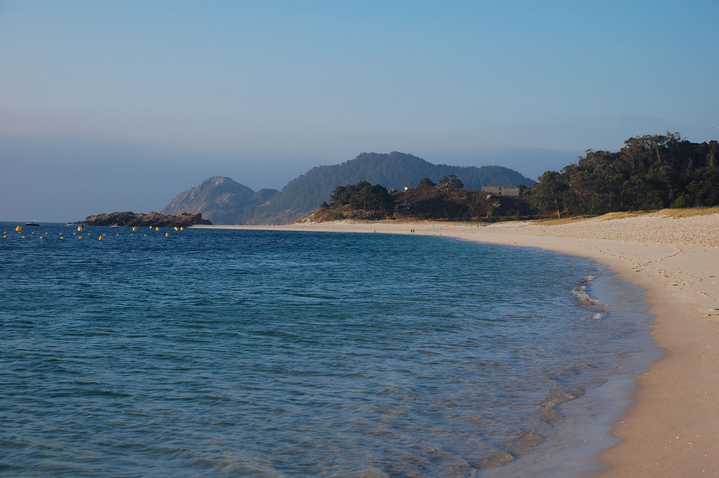 A beach with clear blue water, sandy shore, and a forested hill in the background.
