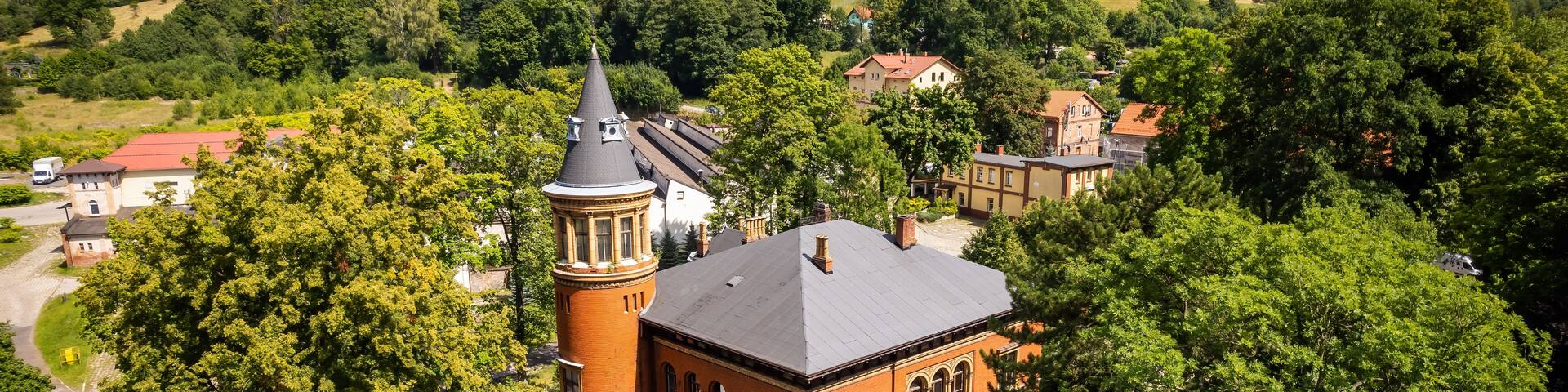 A red brick building with a tower, surrounded by lush green trees and hills in the background.
