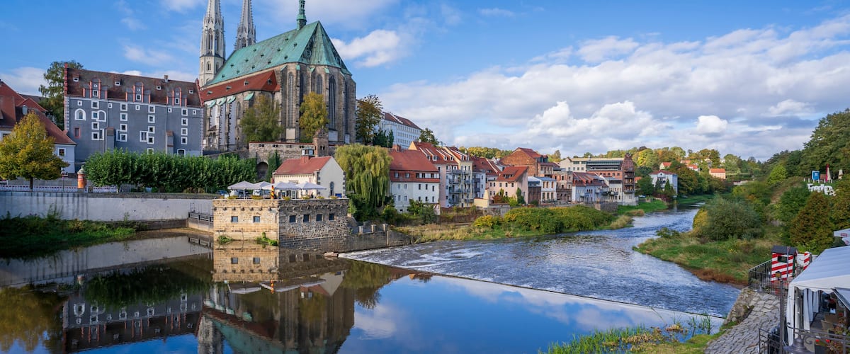 A river reflecting a historic church with twin spires and a green roof, surrounded by traditional buildings and a cobblestone path.