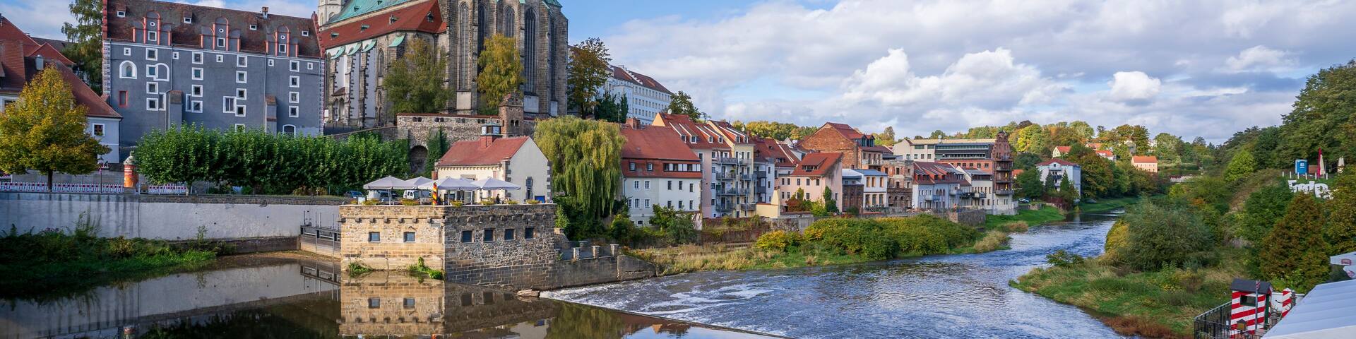 A river reflecting a historic church with twin spires and a green roof, surrounded by traditional buildings and a cobblestone path.