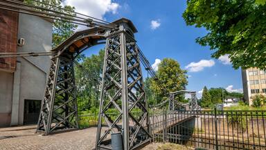 Une structure de pont métallique avec un escalier sur le côté droit, entourée de verdure et avec un bâtiment en arrière-plan.