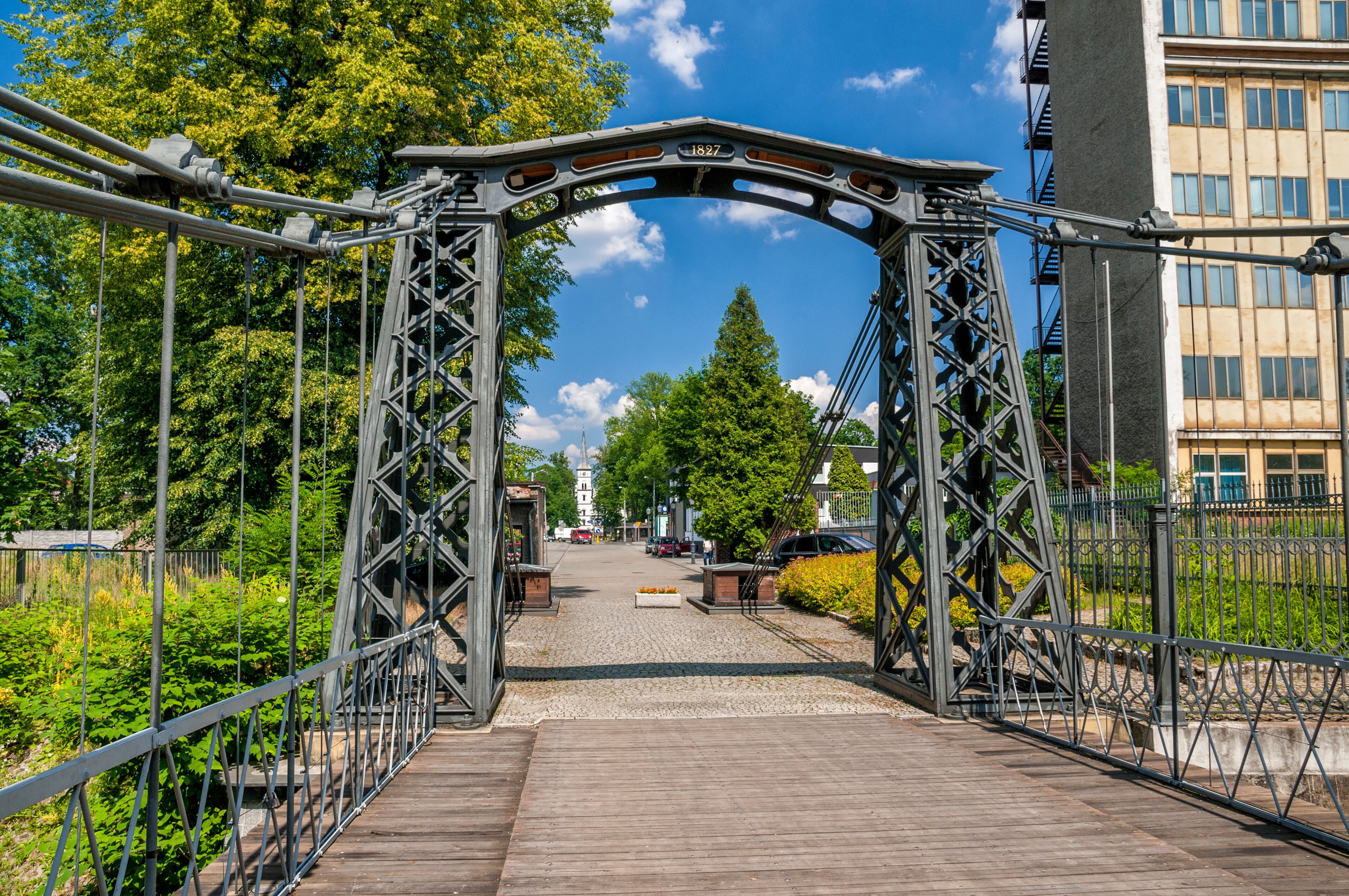 A metal bridge with a date plaque, surrounded by greenery and buildings.