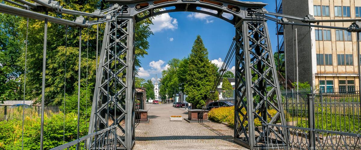 A metal bridge with a date plaque, surrounded by greenery and buildings.