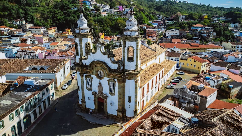 A historic church with two bell towers, surrounded by colorful buildings and a hilly landscape.