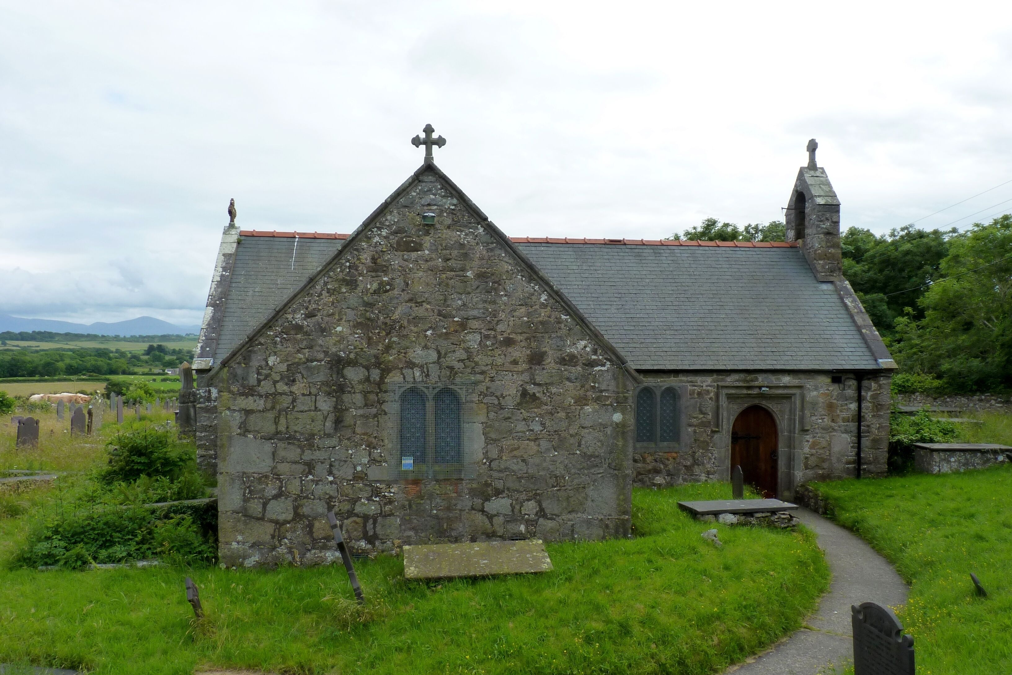 A stone church with a cross on top, surrounded by a cemetery and greenery.