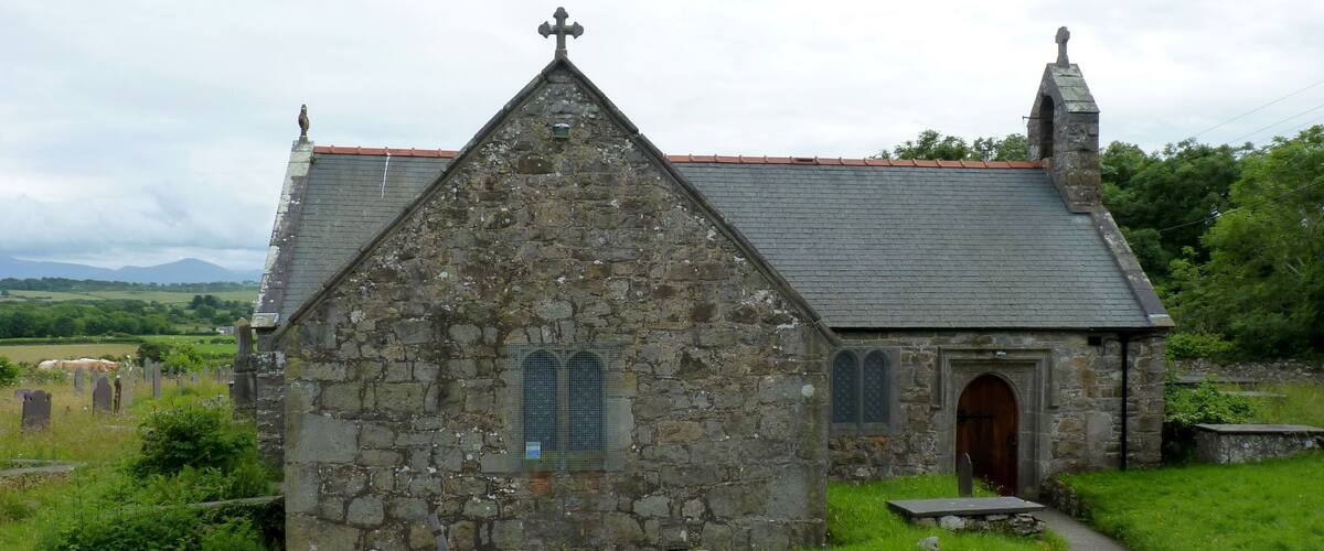 A stone church with a cross on top, surrounded by a cemetery and greenery.