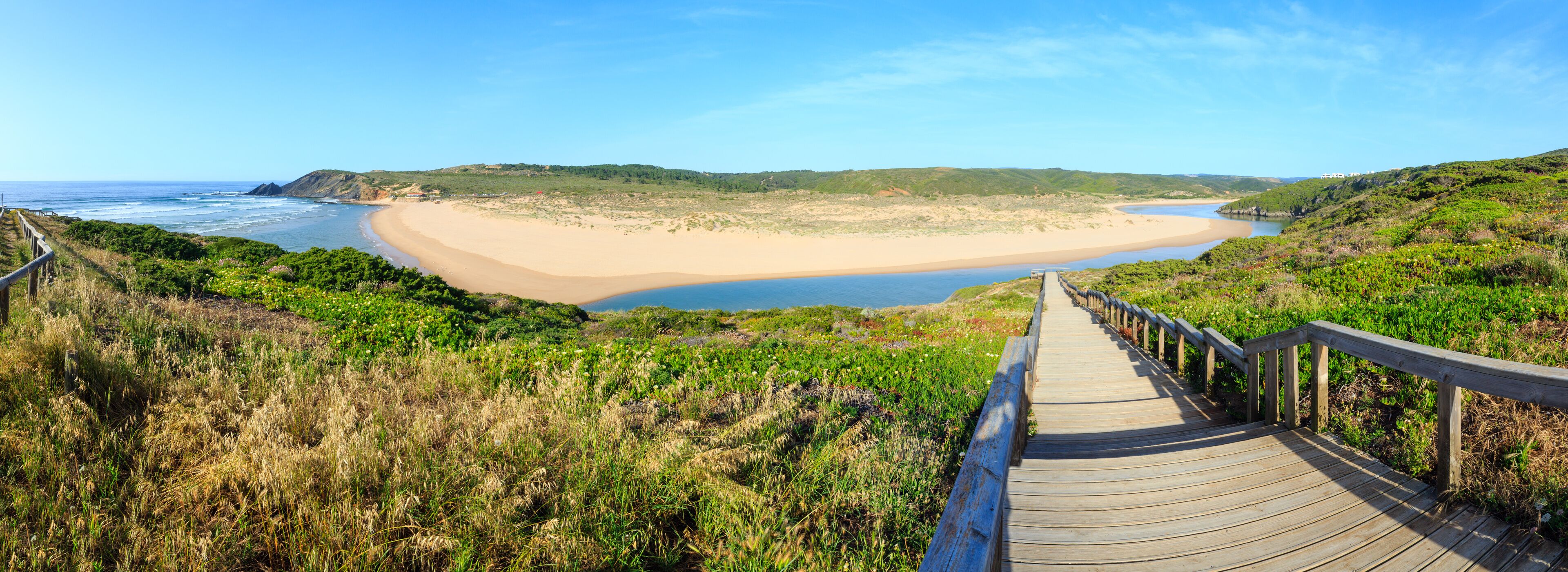 A coastal landscape with a wooden boardwalk leading to a sandy beach and a calm sea.