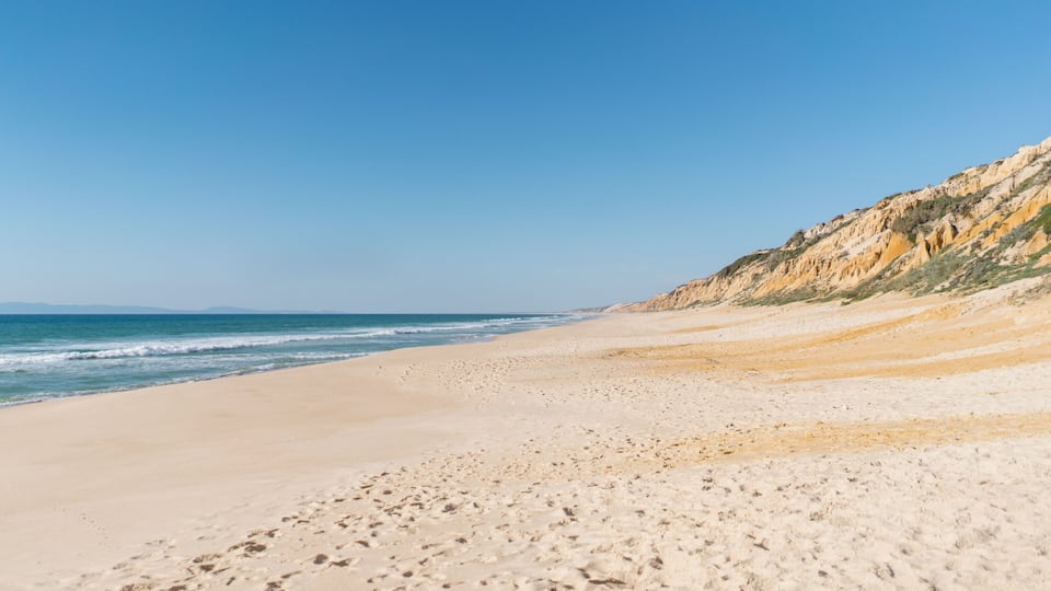 A sandy beach with gentle waves and a rocky cliff in the background.