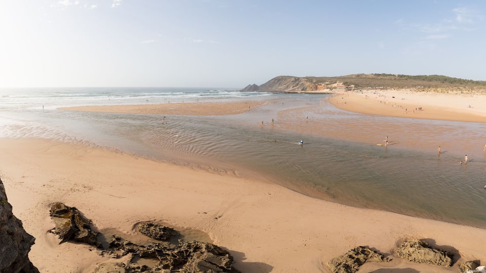 A sandy beach with calm waters, rocky outcrops, and distant hills.