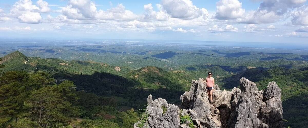 A person standing on rocky outcrops overlooking a vast landscape of green hills and valleys under a partly cloudy sky.