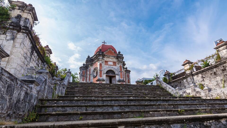 A historic church with a red dome and a cross, surrounded by stone walls and steps leading up to it.