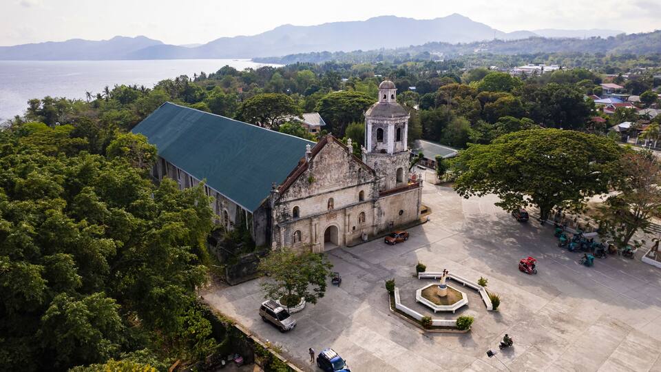 An aerial view of a historic church with a bell tower, surrounded by lush greenery and a body of water in the background.