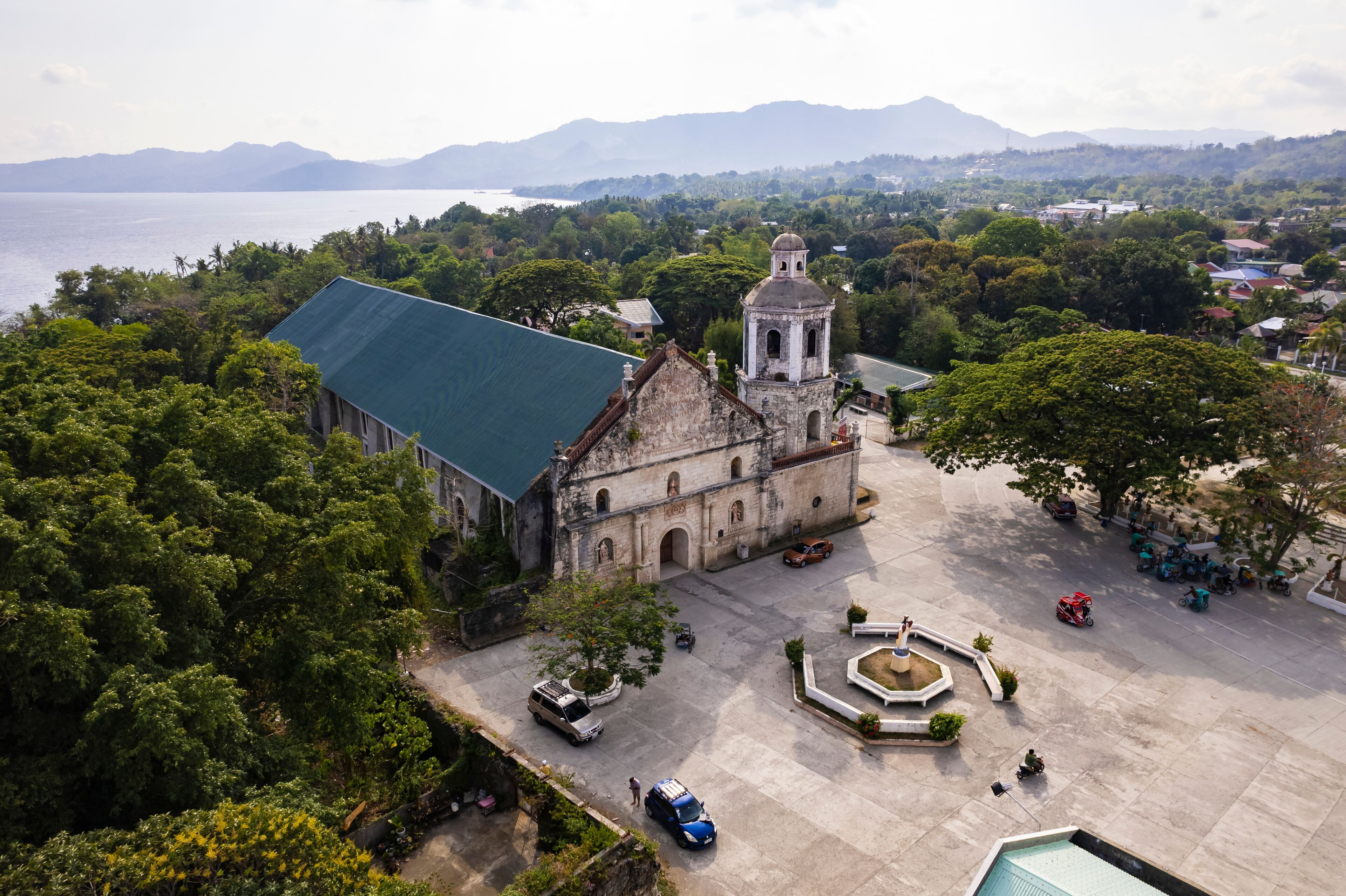 An aerial view of a historic church with a bell tower, surrounded by lush greenery and a body of water in the background.