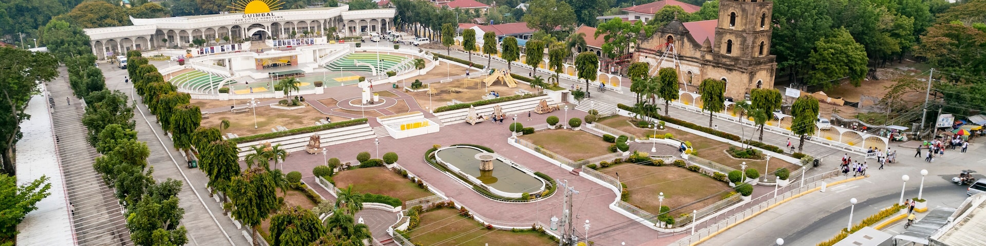 Una amplia plaza abierta con una fuente central, rodeada de árboles y edificios, incluyendo una iglesia con un campanario.