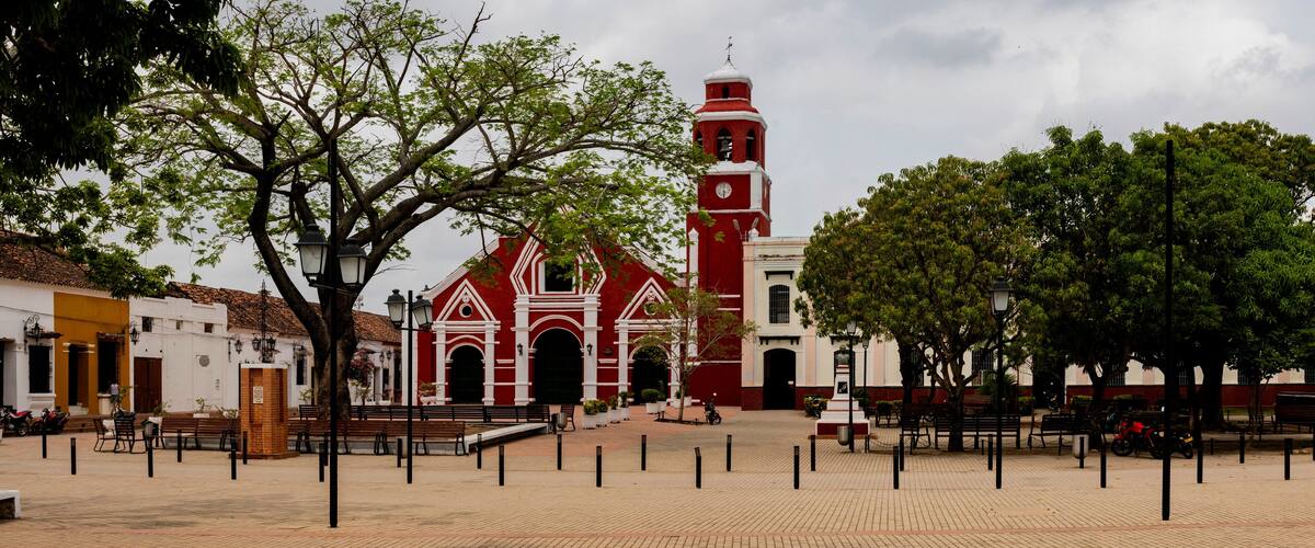 A plaza with a red church, benches, trees, and a white building.