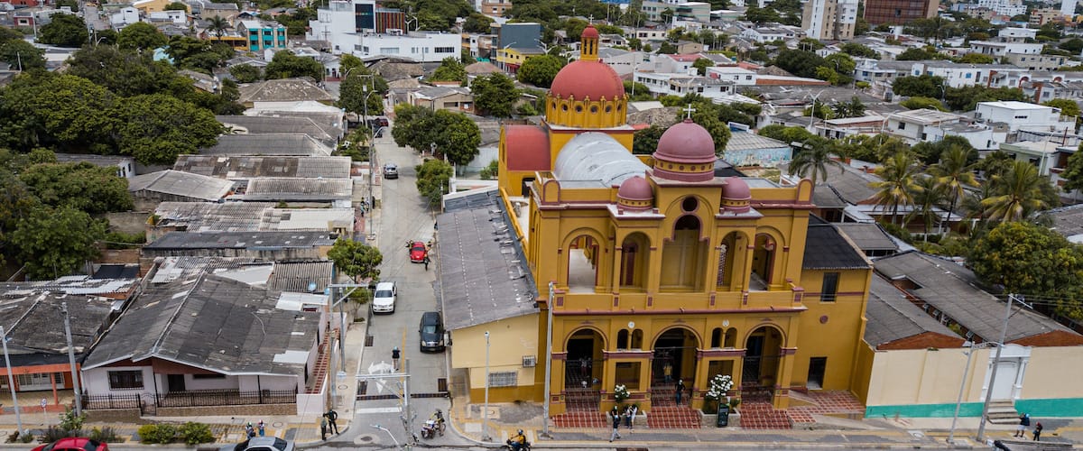 A yellow church with red domes and a white cross, surrounded by residential buildings and palm trees.