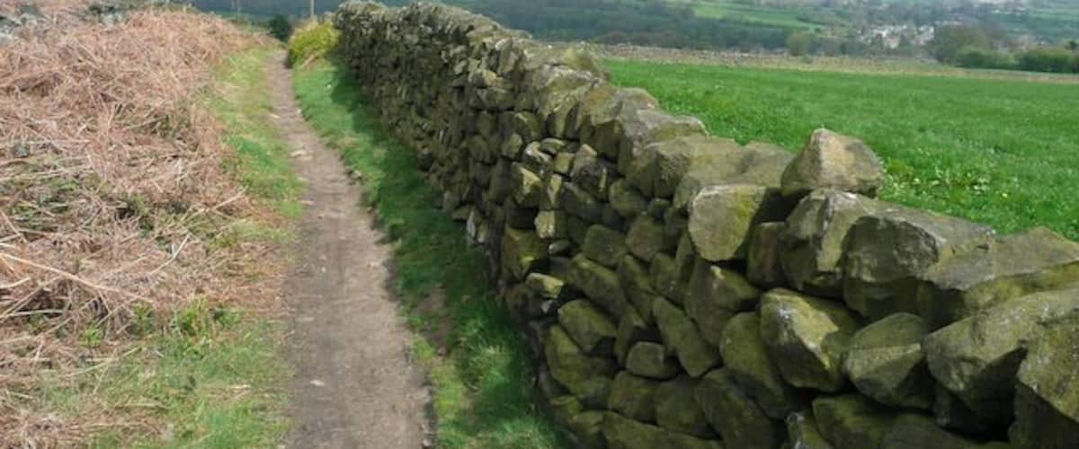 A stone wall beside a narrow path with a rural landscape in the background.
