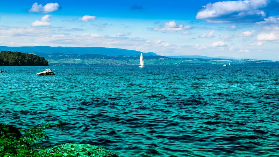 A turquoise sea with a sailboat, rocky shore, and distant mountains.