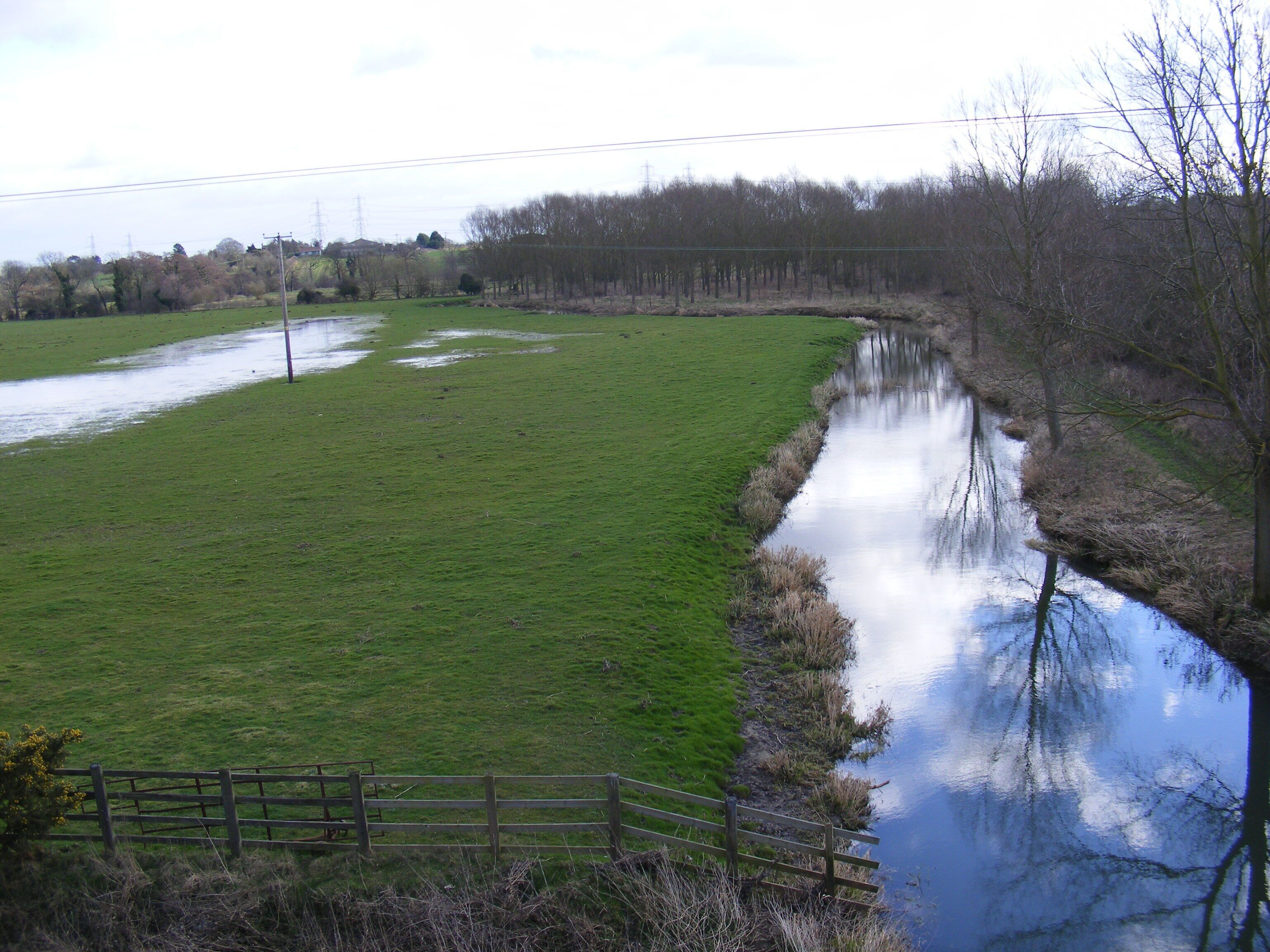 A rural landscape with a river reflecting trees and a grassy field.