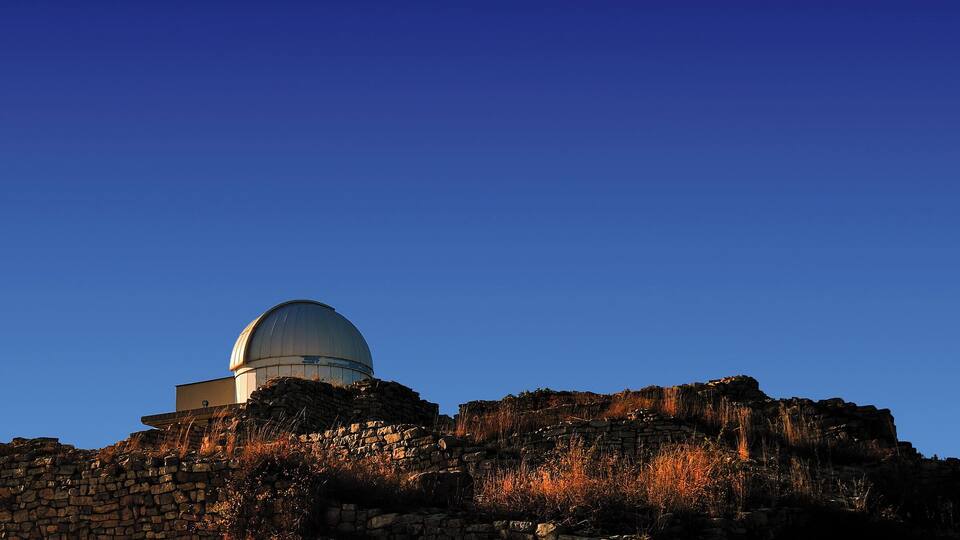 A large observatory dome on a hill with stone walls and dry grass.