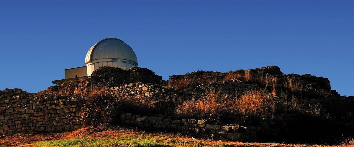 A large observatory dome on a hill with stone walls and dry grass.