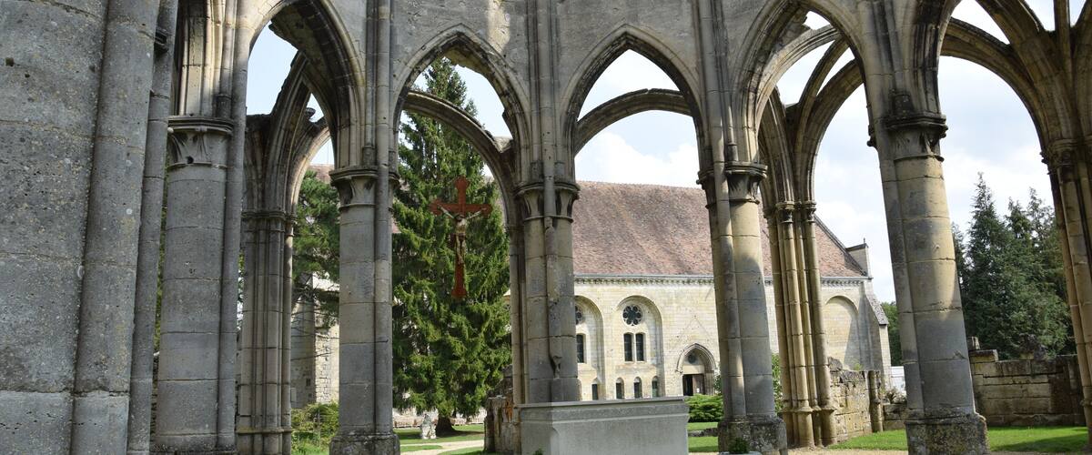 A Gothic-style cloister with stone arches and columns, a central tomb, and a building with a tiled roof in the background.