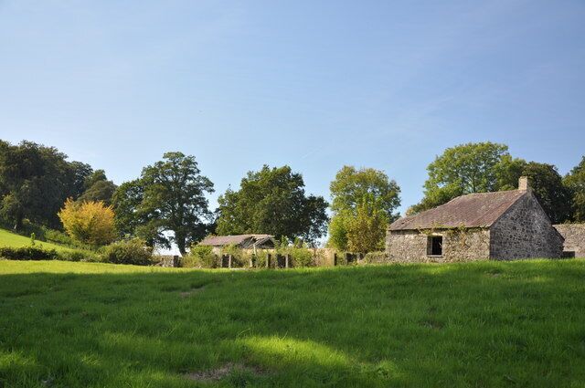 A stone house with a thatched roof in a grassy field, surrounded by trees and a clear blue sky.