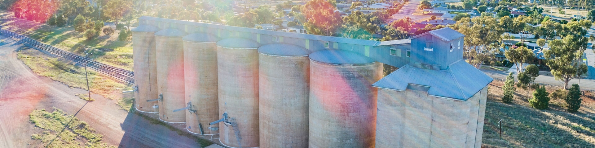 Complejo de silos de grano con varios silos y un pequeño edificio, rodeado de un paisaje rural con árboles y cielo despejado.