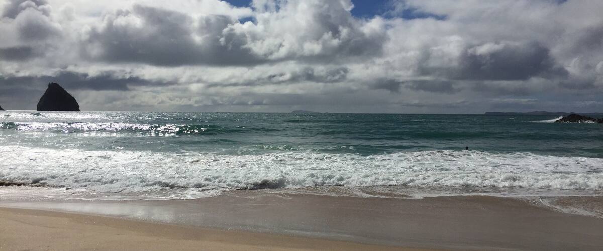 A beach with waves, a large rock formation, and a cloudy sky.