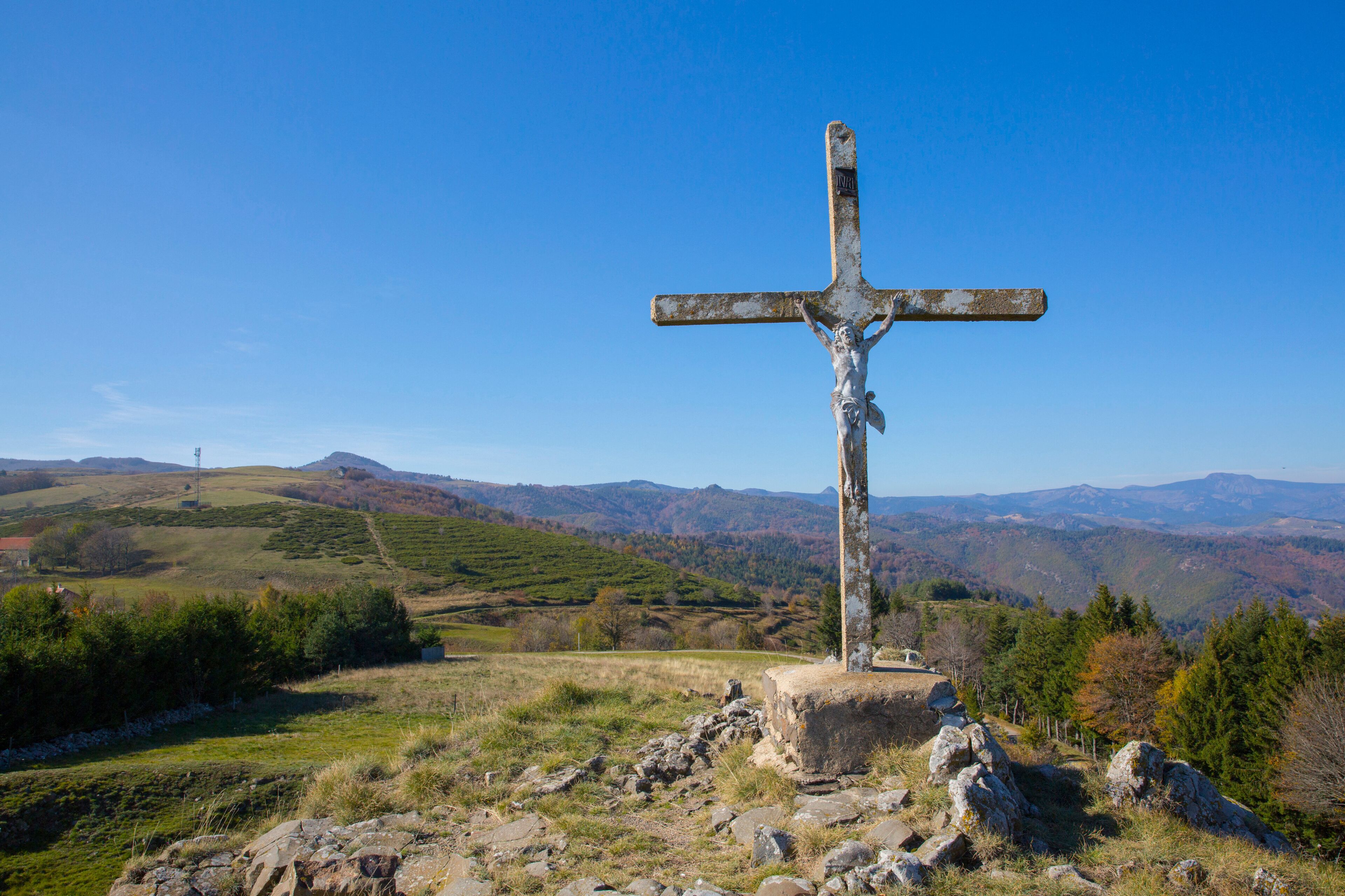 A weathered cross on a hilltop with a scenic view of rolling hills and mountains in the background.