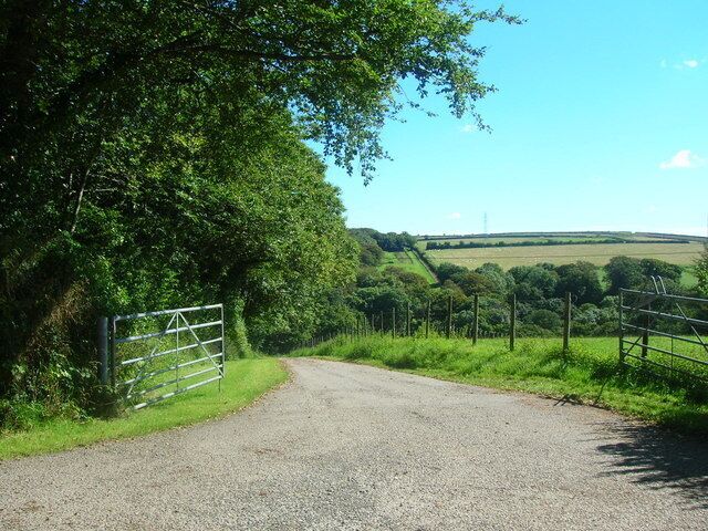 A gravel road leading through a rural area with a metal gate, surrounded by greenery and a clear sky.