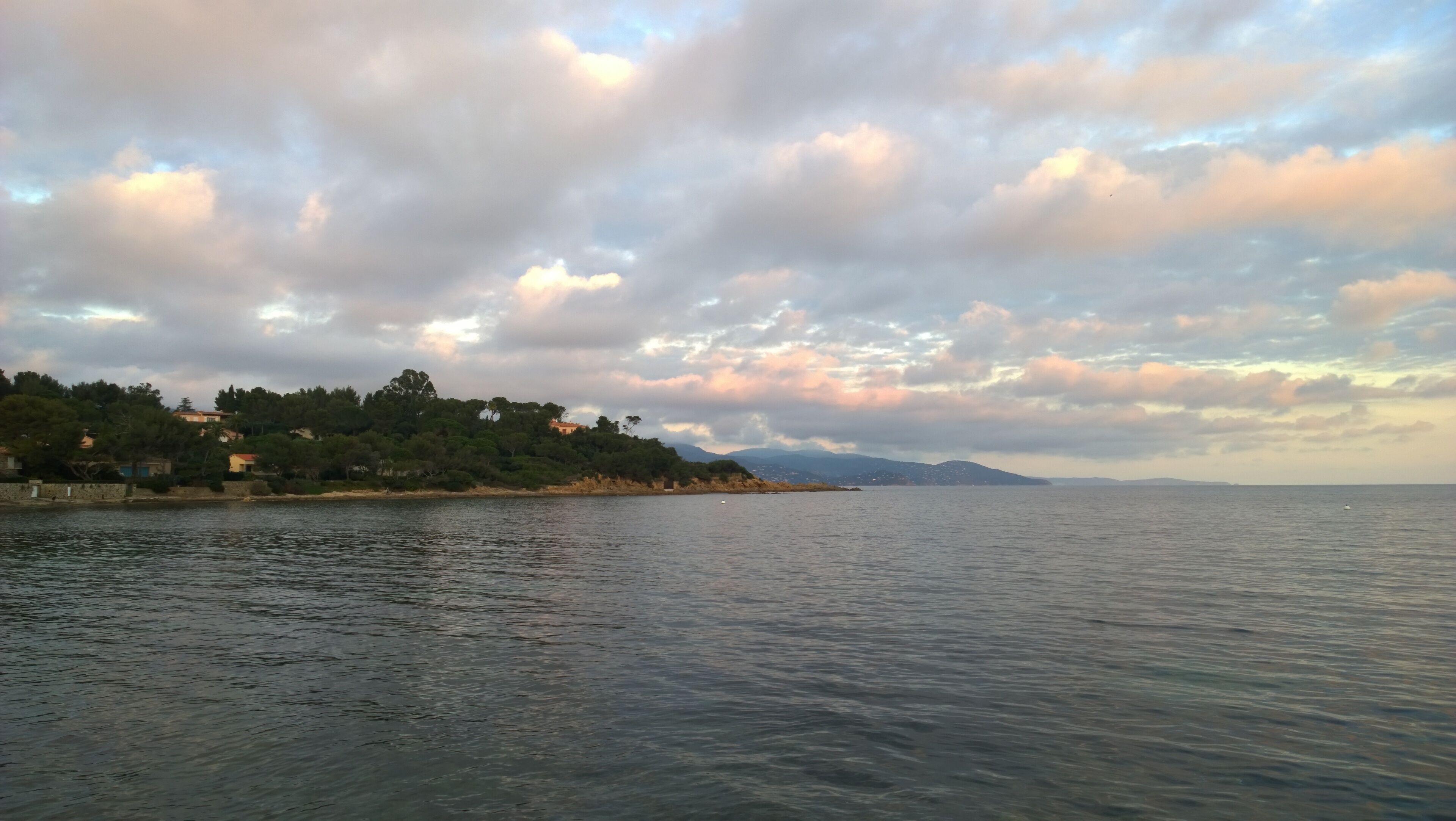 Un paisaje costero con mar tranquilo, una colina cubierta de árboles y montañas lejanas bajo un cielo nublado.