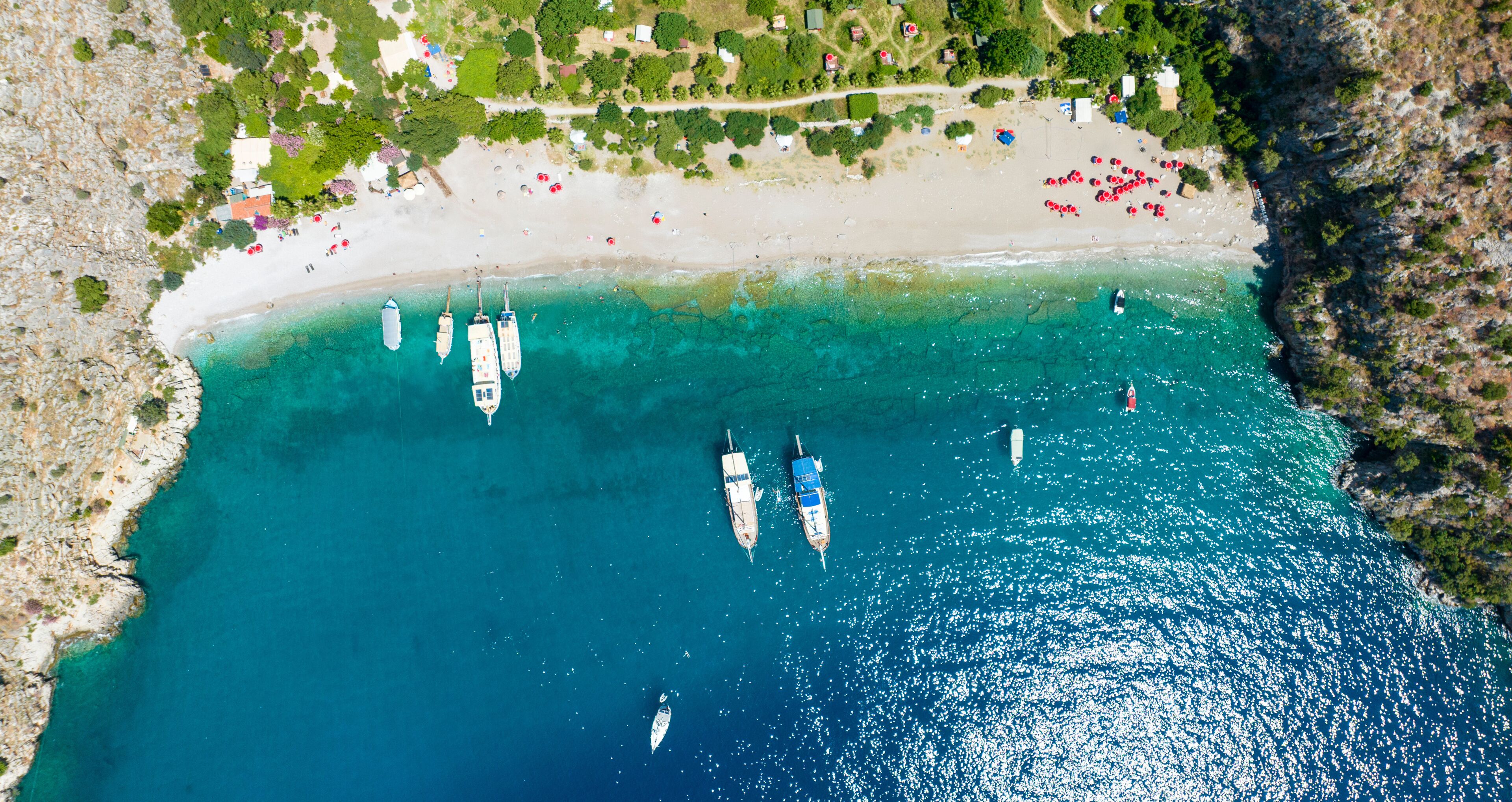 An aerial view of a coastal area with a sandy beach, several boats anchored in the water, and a rocky coastline.