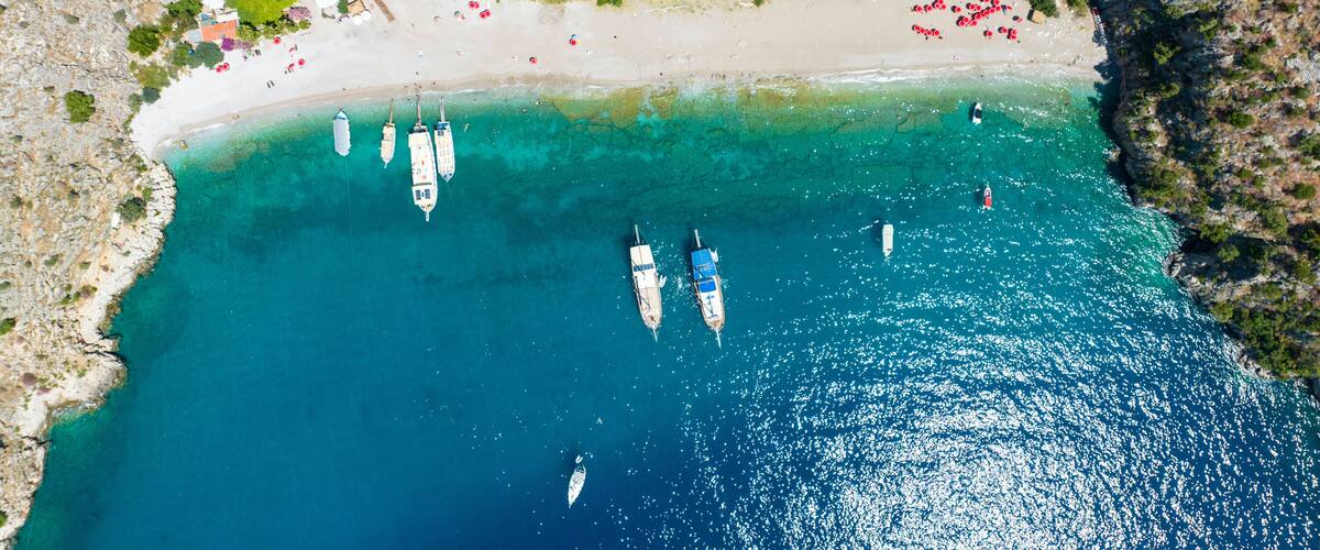 An aerial view of a coastal area with a sandy beach, several boats anchored in the water, and a rocky coastline.
