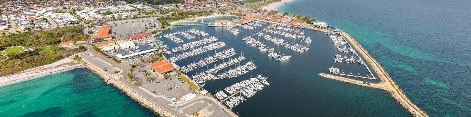 An aerial view of a marina with boats docked, surrounded by a coastal town and a clear sky.