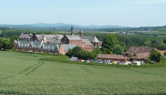 A large red brick building with multiple towers, surrounded by green fields and a few smaller houses.
