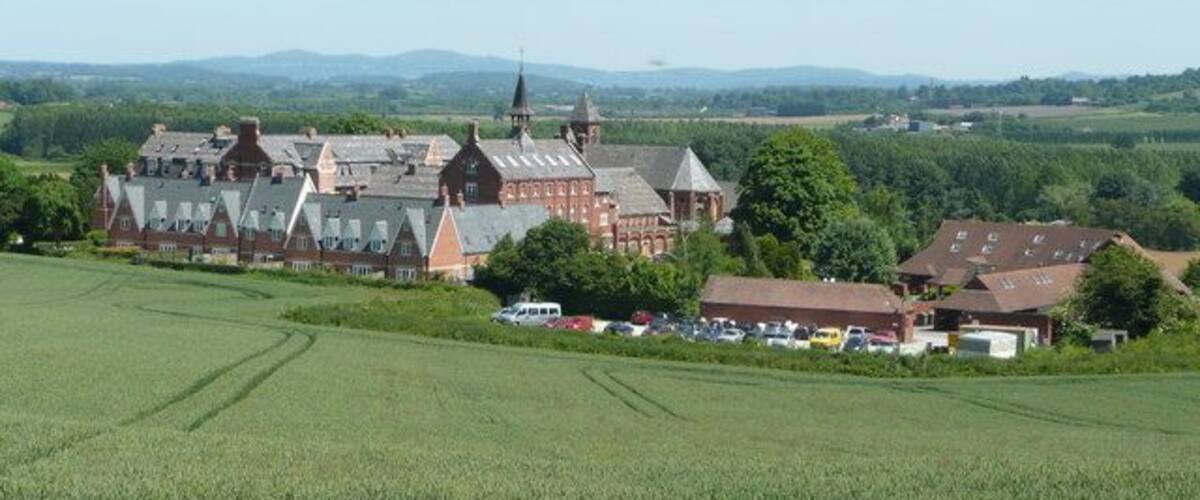 A large red brick building with multiple towers, surrounded by green fields and a few smaller houses.