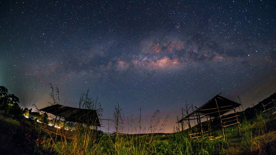 A starry night sky with the Milky Way visible, a rural landscape with two wooden structures, and tall grass in the foreground.