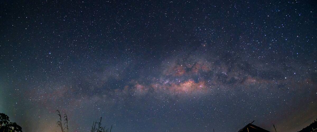 A starry night sky with the Milky Way visible, a rural landscape with two wooden structures, and tall grass in the foreground.