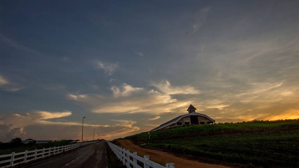 A dirt road with a white fence, a barn, and a cloudy sky.
