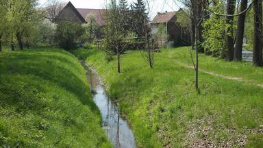 Ein schmaler Fluss schlängelt sich durch eine üppige, grüne Landschaft mit Bäumen und einem Steinhaus im Hintergrund.