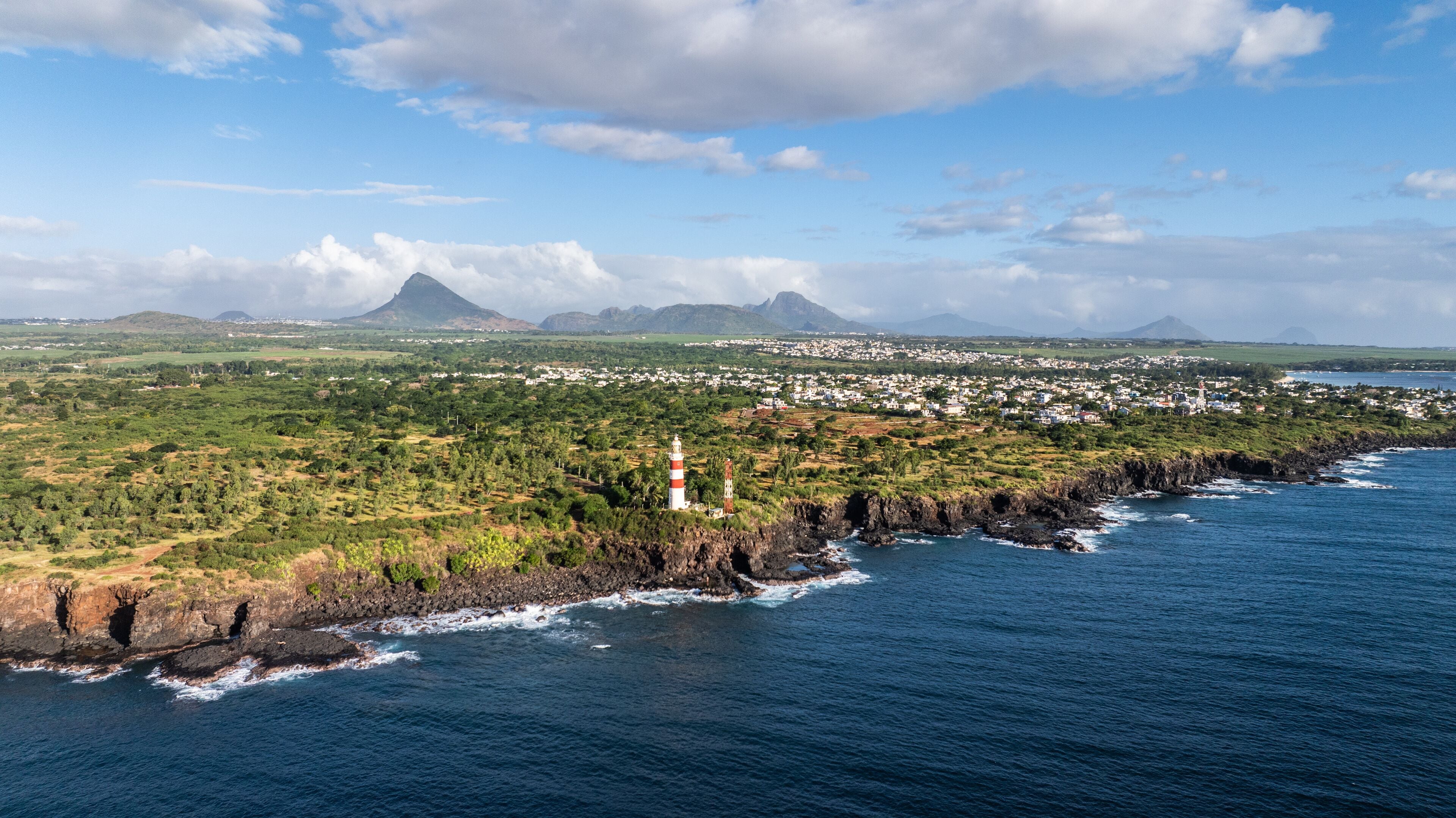 A coastal town with a lighthouse, green hills, and a volcano in the background.