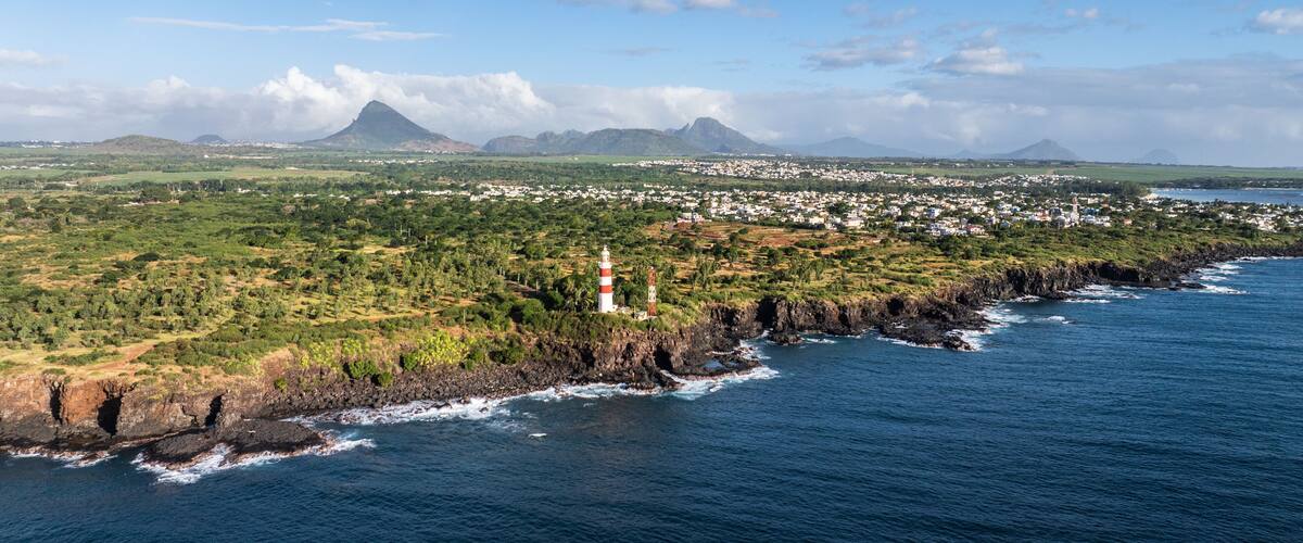 A coastal town with a lighthouse, green hills, and a volcano in the background.