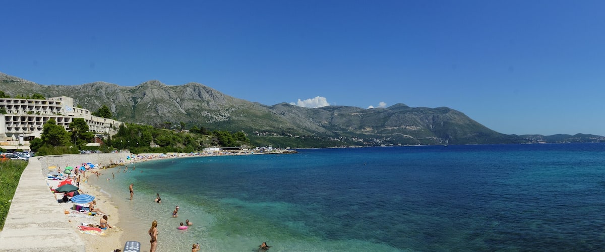 A coastal town with a beach, clear blue sea, mountains, and a modern building.