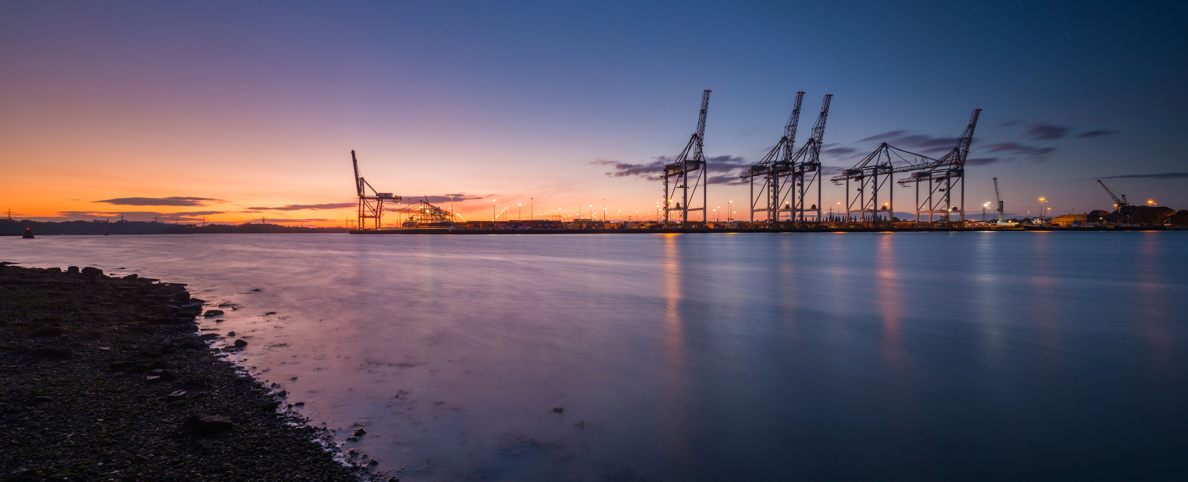 A calm waterfront at dusk with industrial cranes and a clear sky.