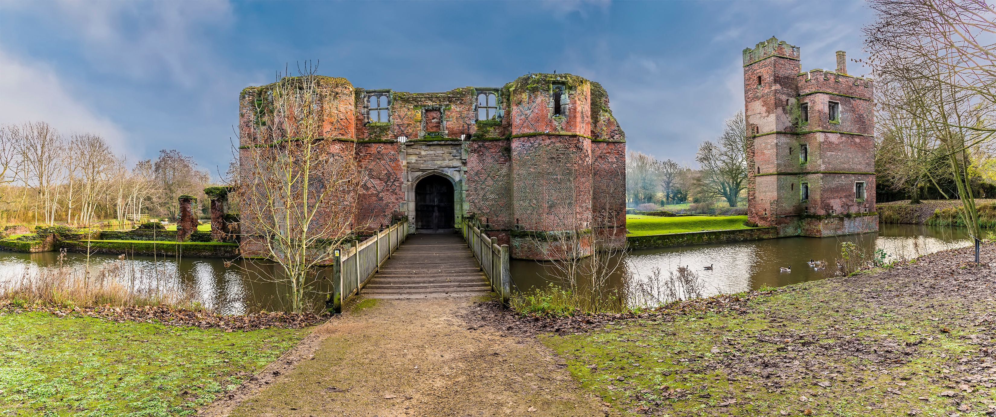A historic castle with a moat, a bridge leading to the entrance, and a serene parkland surrounding it.