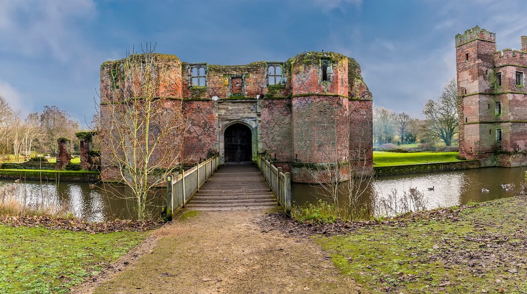 A historic castle with a moat, a bridge leading to the entrance, and a serene parkland surrounding it.