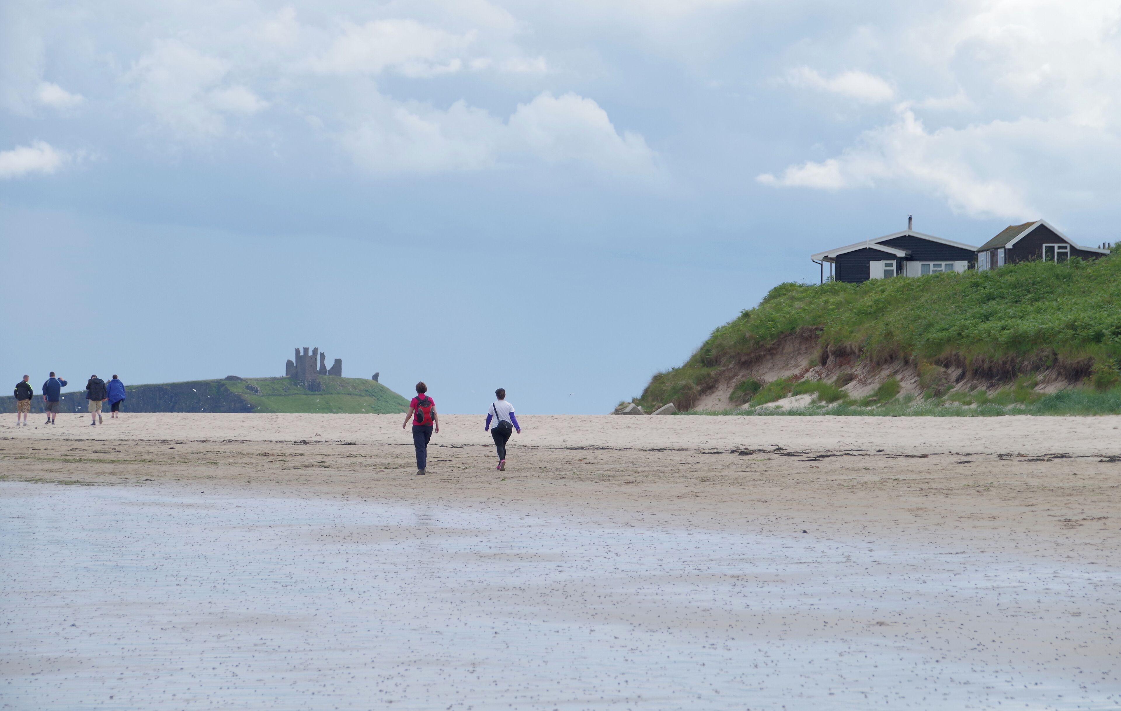 A beach with people walking, a grassy hill with a house, and a distant castle.