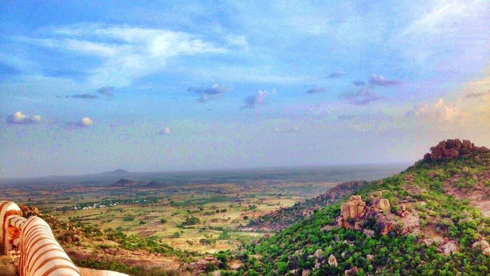 A landscape with a large rock formation, green vegetation, and a distant view of a valley.