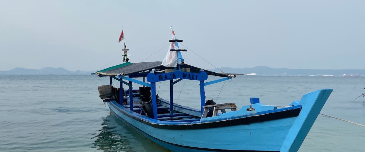 A blue boat named "RAJA WALI" anchored in calm waters with a clear sky.